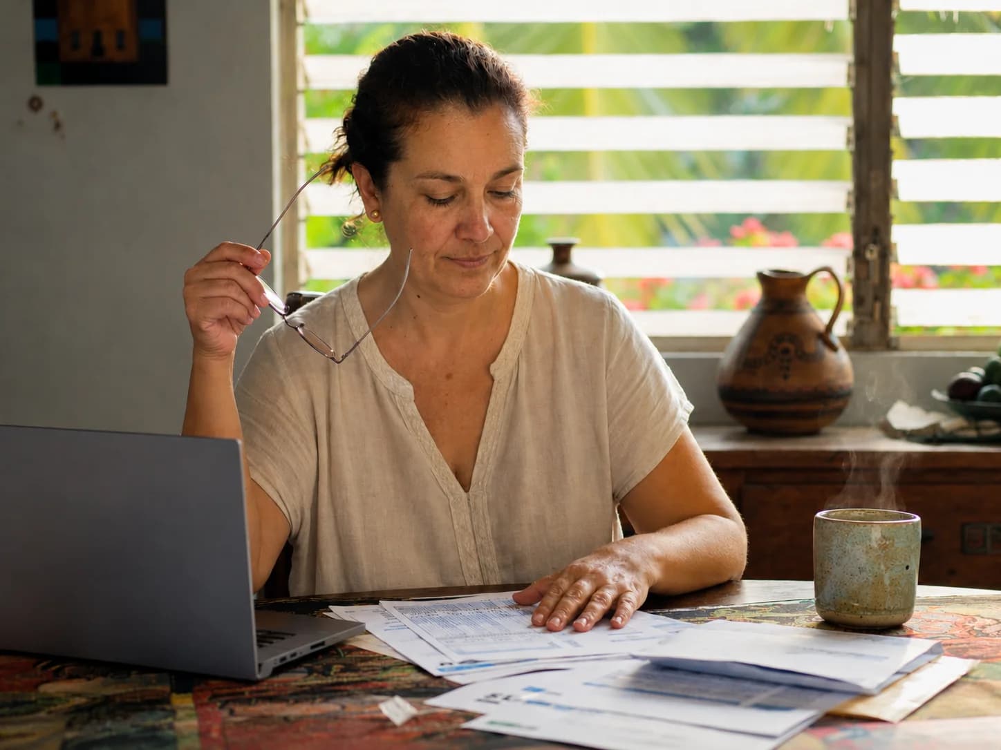 Mujer costarricense de unos 48 años revisando documentos de seguro médico y CCSS en la mesa de su cocina, con anteojos de lectura en la mano y un café humeante junto a la laptop.